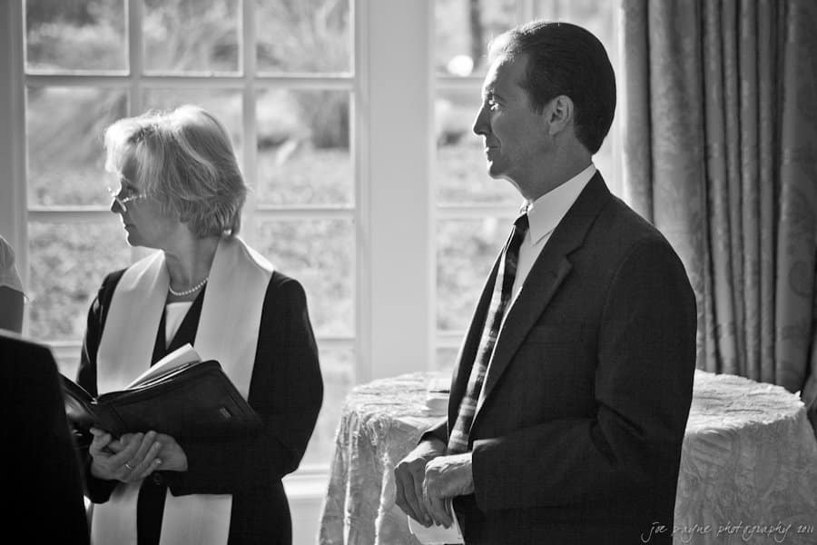 A black and white image of a woman in a robe holding an open book and a man in a suit standing side by side. They are indoors, near a window with sheer curtains, and appear to be part of a formal ceremony or event.