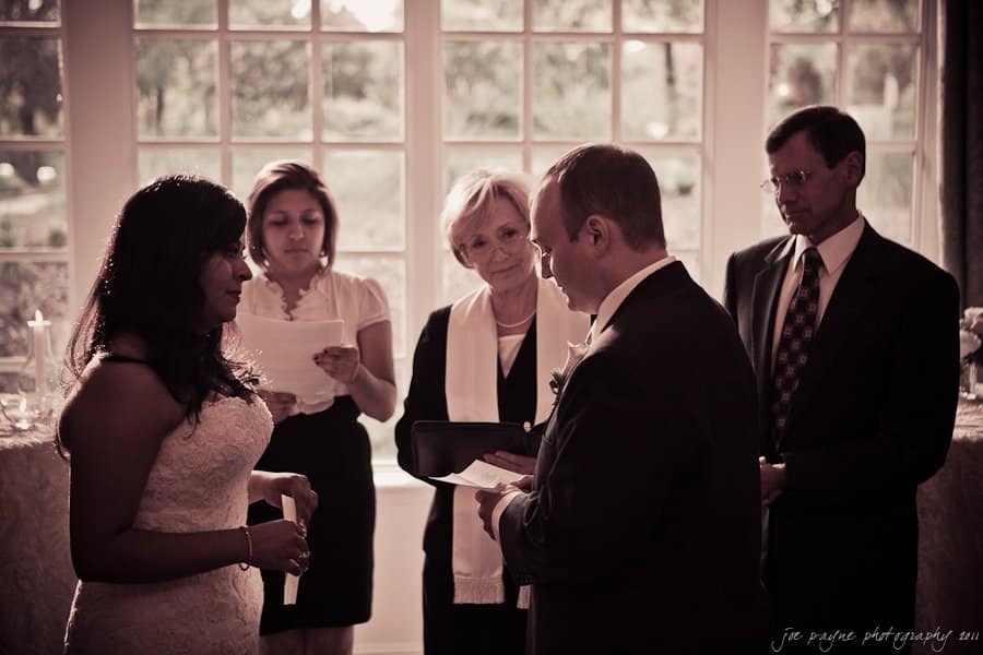 A couple stands facing each other during a wedding ceremony. The bride wears a white dress, and the groom is in a suit. An officiant and two witnesses are present, with a large window and outdoor scenery in the background.