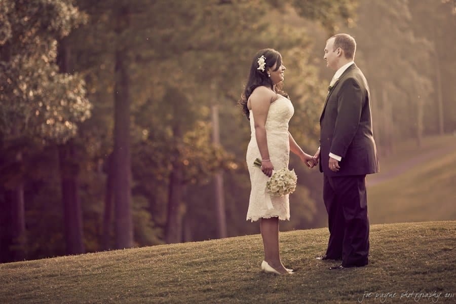 A bride and groom stand facing each other outdoors at the Washington Duke Inn, holding hands and smiling, surrounded by trees and soft sunlight, with the bride holding a bouquet—a perfect moment captured by Durham wedding photography.