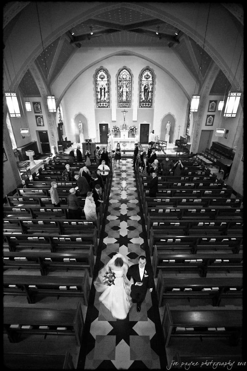 A bride and groom walk down the aisle of a church with vaulted ceilings and stained glass windows. Guests stand and watch, while some are seated in wooden pews. The floor features a patterned design, and the altar is adorned with decorations.