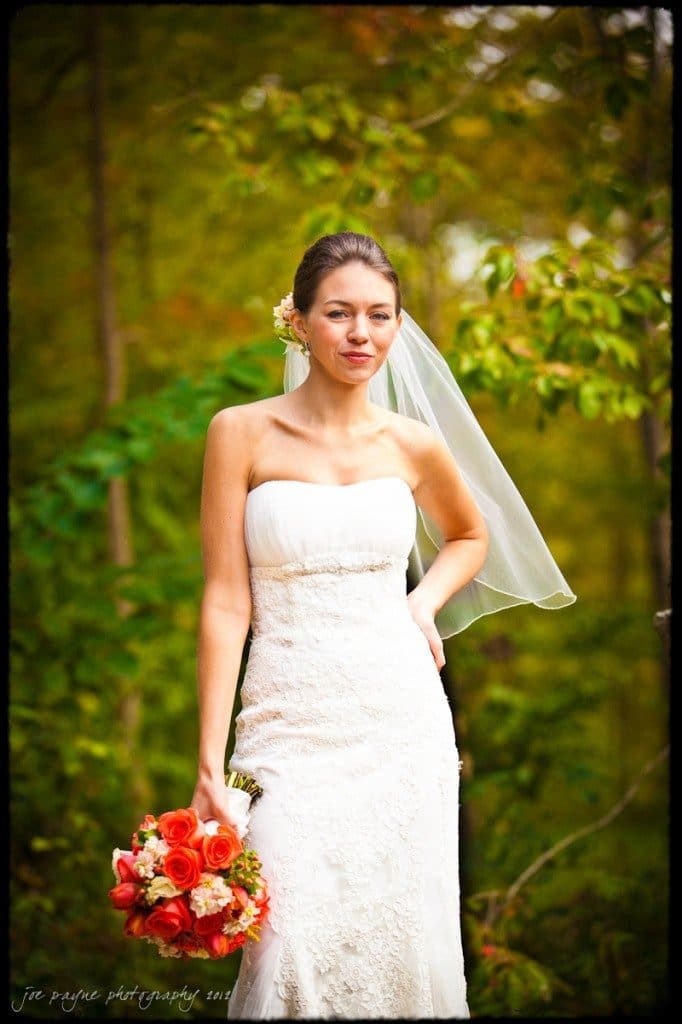 A bride in a strapless white wedding dress stands outdoors with a bouquet of red and orange flowers. She has a veil and a flower in her hair. Lush greenery surrounds her.