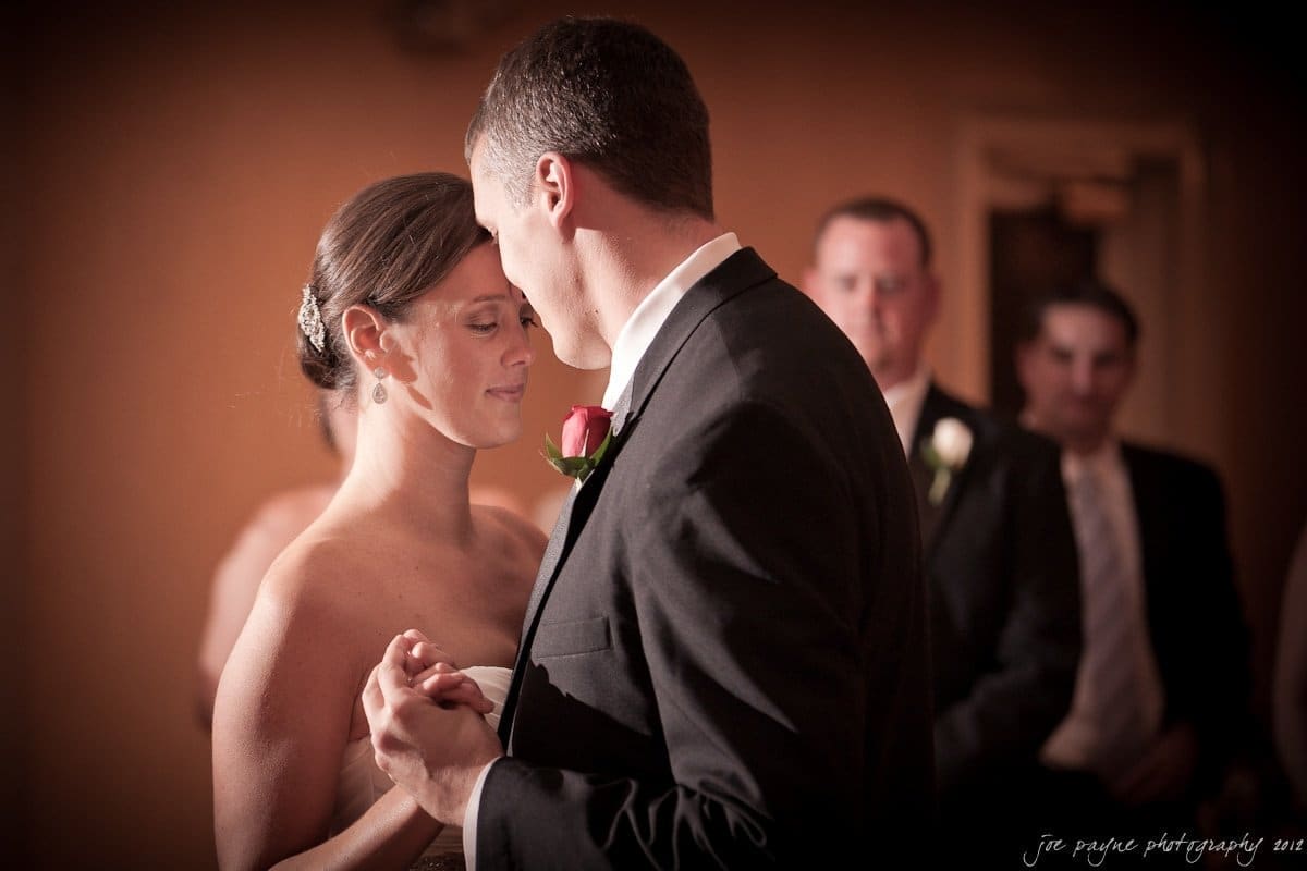 A bride and groom share a dance in a dimly lit room. The bride, with her hair elegantly styled, wears a white dress, and the groom is in a black suit with a red rose boutonniere. They hold hands closely, surrounded by blurred guests in the background.