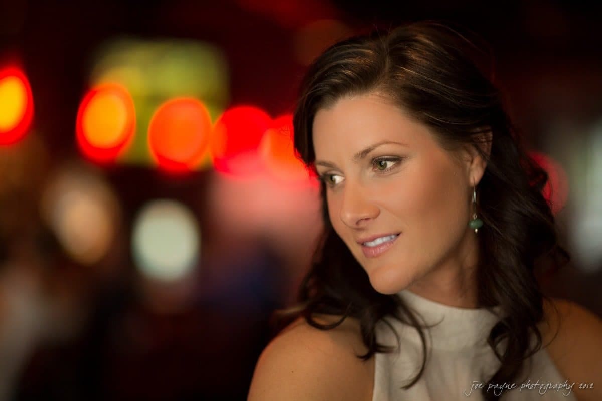 downtown raleigh engagement session ~ lisa and mark A woman with long dark hair and a sleeveless top gazes to the side. The background is softly blurred with warm, colorful lights creating a bokeh effect.