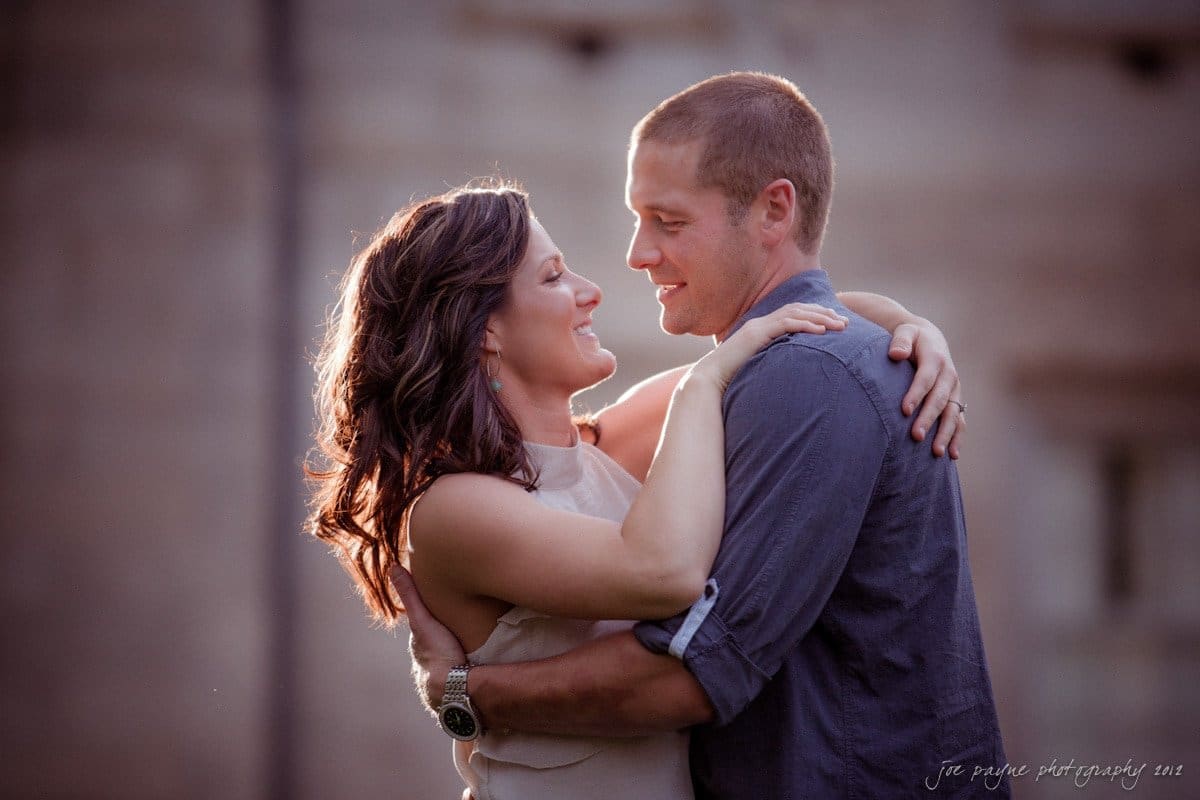 downtown raleigh engagement session ~ lisa and mark A couple embraces lovingly, standing close and smiling at each other. The woman has long brown hair and is wearing a sleeveless beige top, while the man has short hair and is wearing a gray shirt. The background is softly blurred.