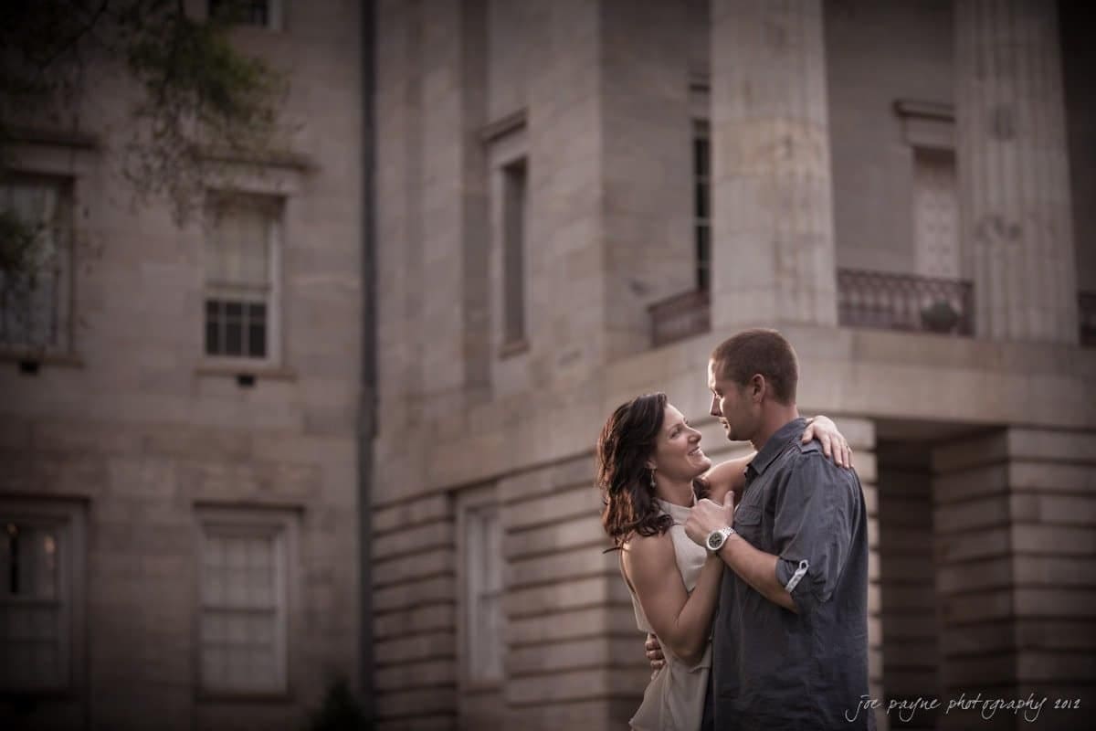 downtown raleigh engagement session ~ lisa and mark A couple embraces and smiles at each other romantically in front of a large, historic stone building with columns. The scene is warmly lit, creating a serene and intimate atmosphere.