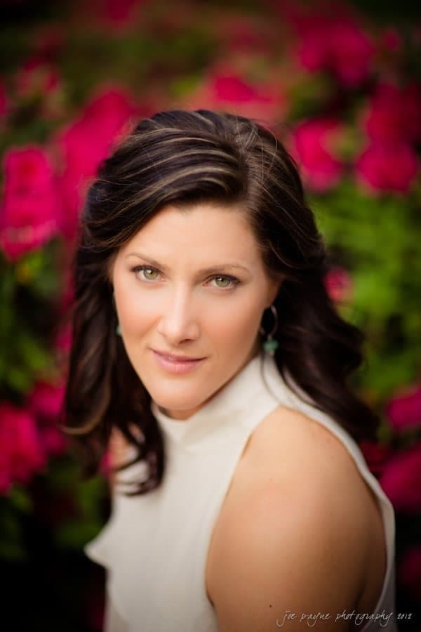 downtown raleigh engagement session ~ lisa and mark A woman with long dark hair and turquoise earrings is wearing a white sleeveless top. She is posing outdoors with vibrant pink flowers and lush green foliage in the background.