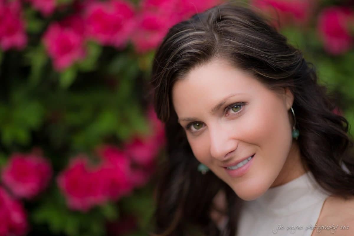 downtown raleigh engagement session ~ lisa and mark A woman with long brown hair smiles at the camera in front of a blurred background of vibrant pink flowers. Shes wearing a light-colored top and turquoise earrings.