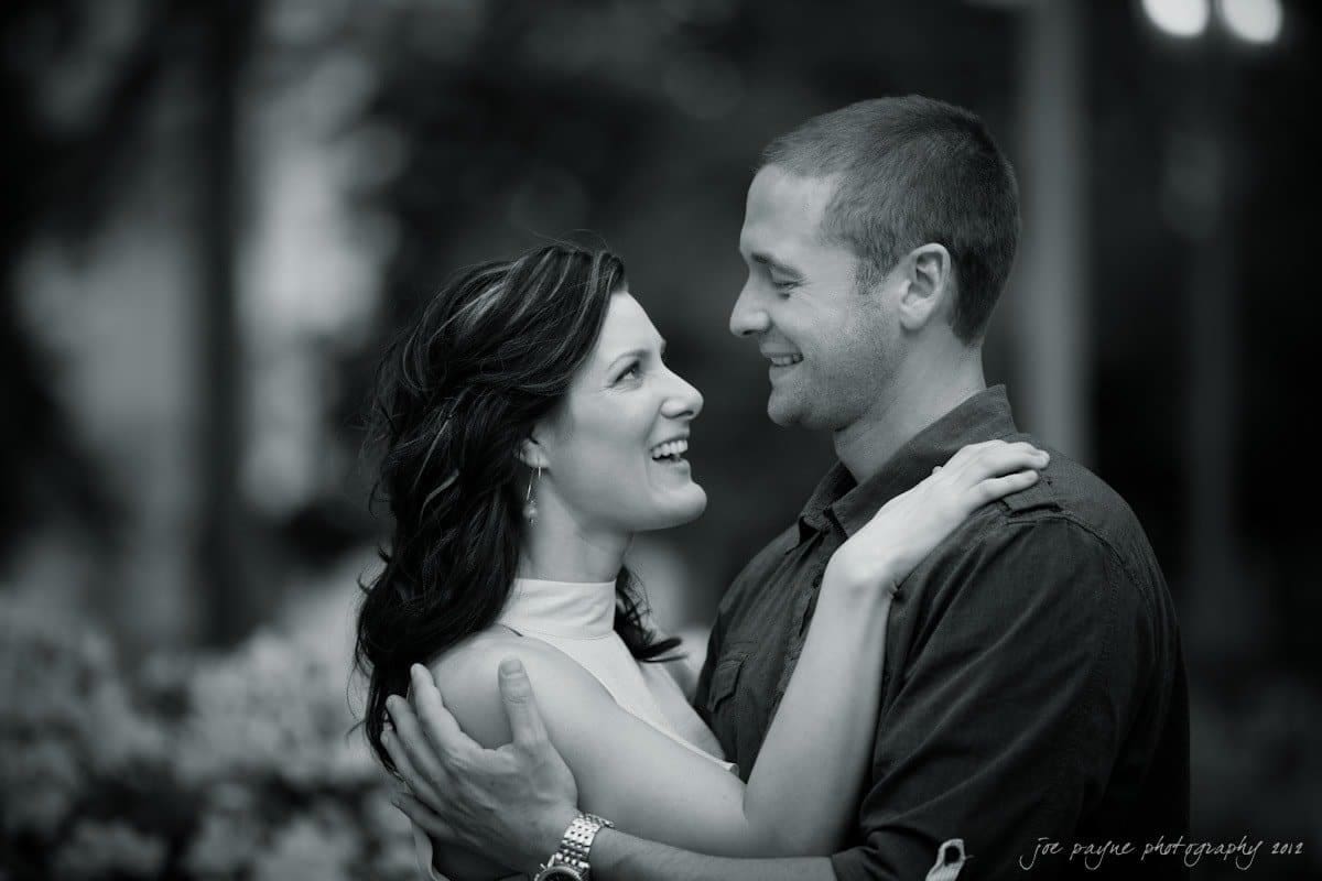 downtown raleigh engagement session ~ lisa and mark A black and white photo of a couple embracing outdoors. They are smiling and looking into each others eyes, with a blurry background of trees and flowers. The woman has long dark hair and is wearing a sleeveless top, while the man is in a shirt.