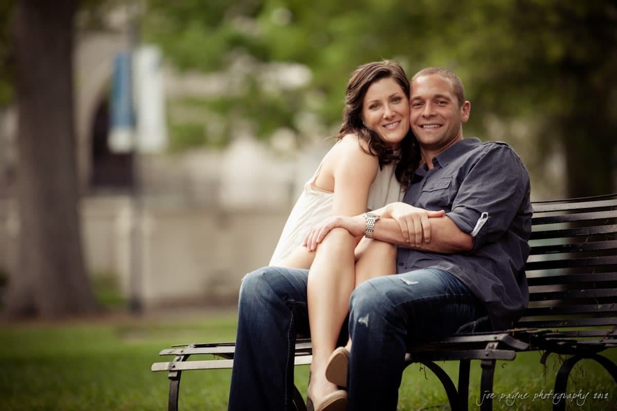 downtown raleigh engagement session ~ lisa and mark A couple sits closely on a park bench. The woman leans affectionately on the mans shoulder, smiling. They are surrounded by greenery. The man wears a gray shirt and jeans, the woman a sleeveless white dress.