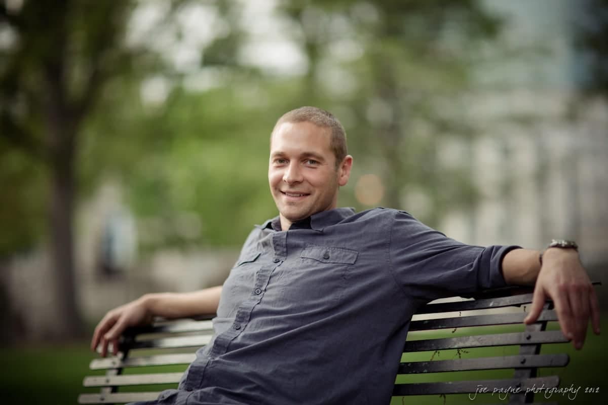 downtown raleigh engagement session ~ lisa and mark A man wearing a gray button-up shirt sits relaxed on a park bench. The background is a blurred view of green trees and grass, suggesting a peaceful outdoor setting.