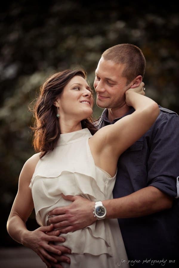 downtown raleigh engagement session ~ lisa and mark A couple embraces outdoors, with the woman facing the camera and the man holding her from behind. They look at each other affectionately, surrounded by a blurred natural background.