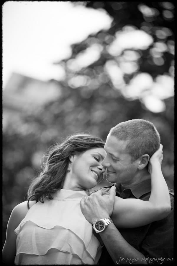 downtown raleigh engagement session ~ lisa and mark A black-and-white photo of a smiling couple embracing outdoors. The woman is leaning into the man, with her eyes closed and a joyful expression. The man is looking at her while gently holding her. Trees are blurred in the background.