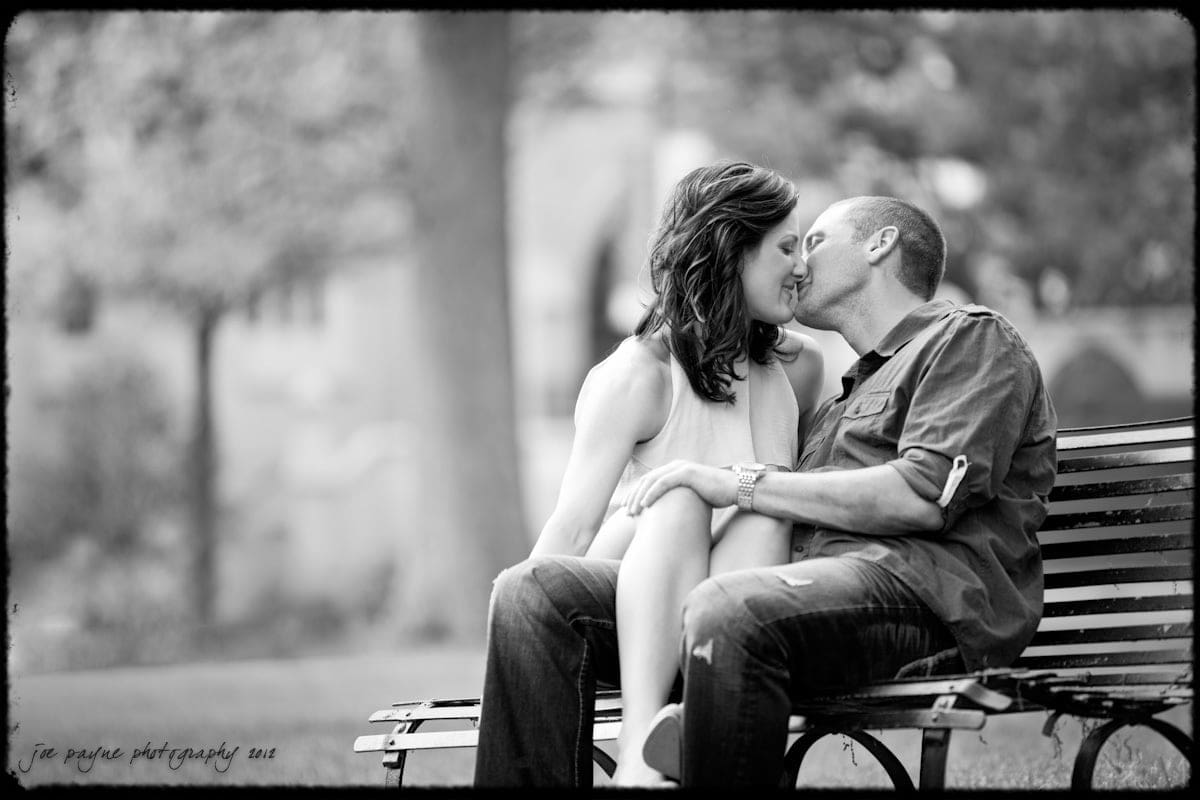 downtown raleigh engagement session ~ lisa and mark A black and white photo of a couple sitting on a park bench, sharing a kiss. The background is blurred, focusing attention on the couples intimate moment. The woman is wearing a sleeveless dress, and the man is in a casual shirt and jeans.