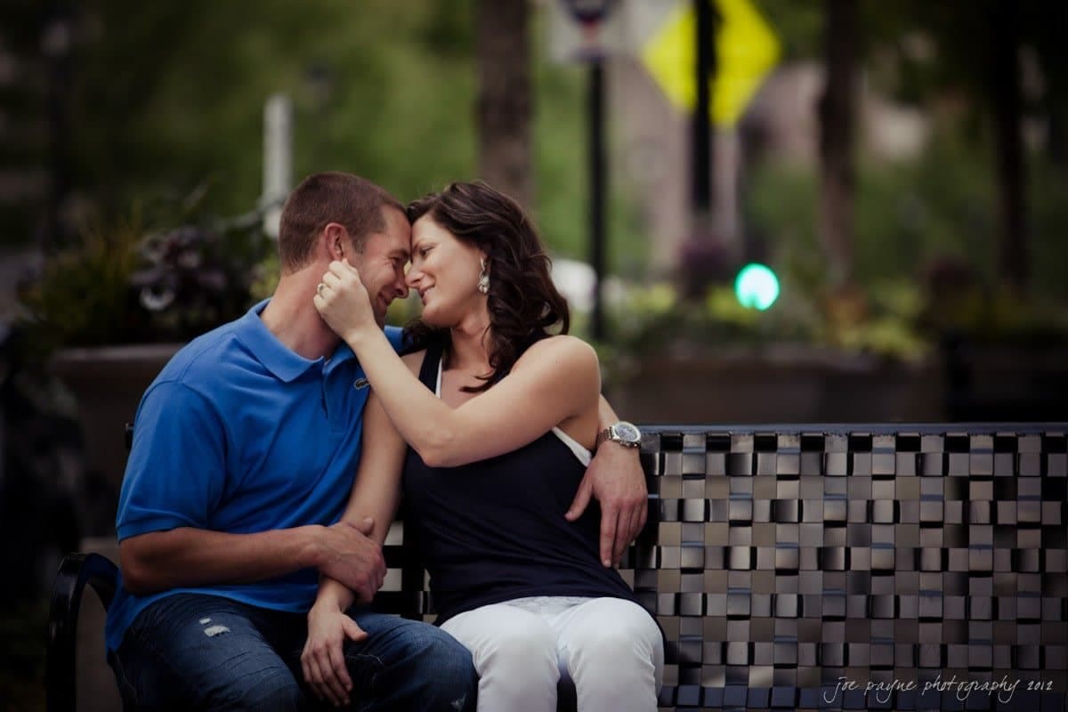 downtown raleigh engagement session ~ lisa and mark A couple sits closely on a park bench, smiling and touching foreheads affectionately. The man wears a blue shirt, and the woman is in a black top with white pants. The background is blurred greenery with soft lighting.