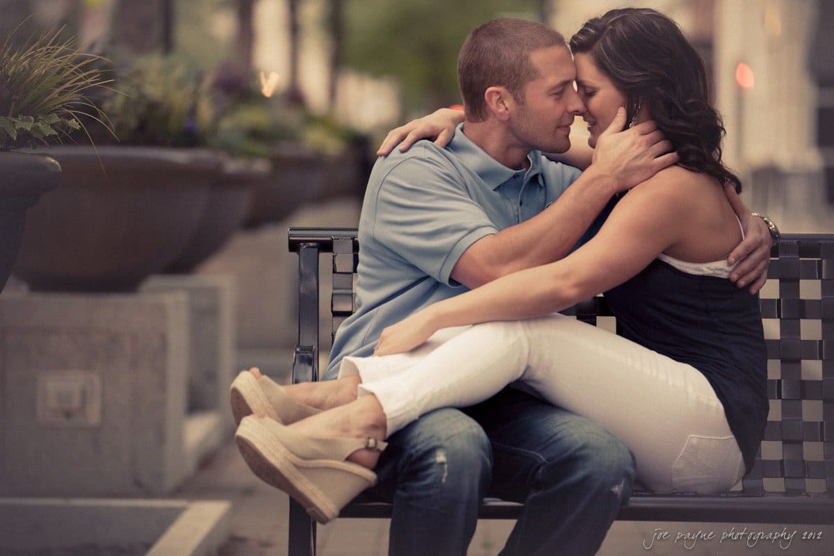 downtown raleigh engagement session ~ lisa and mark A couple sits closely on a bench outdoors, gazing into each others eyes. The woman, wearing white pants and sandals, rests on the mans lap as he gently holds her face. The setting is serene with blurred greenery in the background.