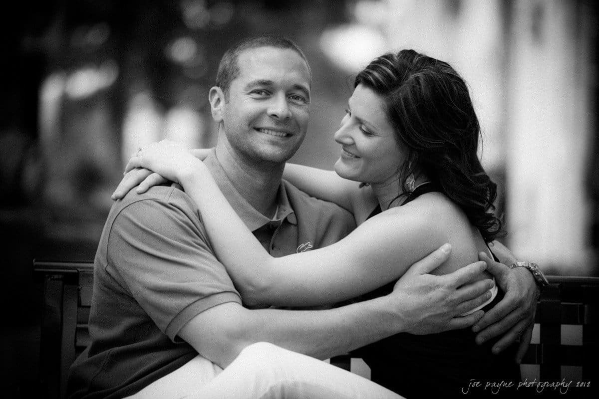 downtown raleigh engagement session ~ lisa and mark Couple sitting closely on a bench, smiling at each other. The man is wearing a polo shirt, and the woman is wearing a sleeveless top. The image is in black and white, capturing a warm and affectionate moment.