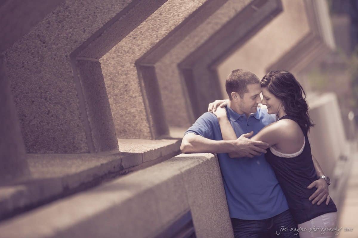 downtown raleigh engagement session ~ lisa and mark A couple embraces, smiling and leaning against a wall with geometric patterns. They wear casual clothing, and the scene has a warm tone, creating a romantic atmosphere.