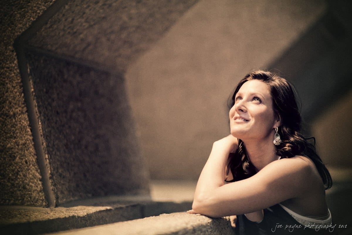 downtown raleigh engagement session ~ lisa and mark A woman with long hair and earrings leans on a textured, angular surface, looking upwards with a smile. The image has a warm, soft focus, creating a contemplative and serene atmosphere.