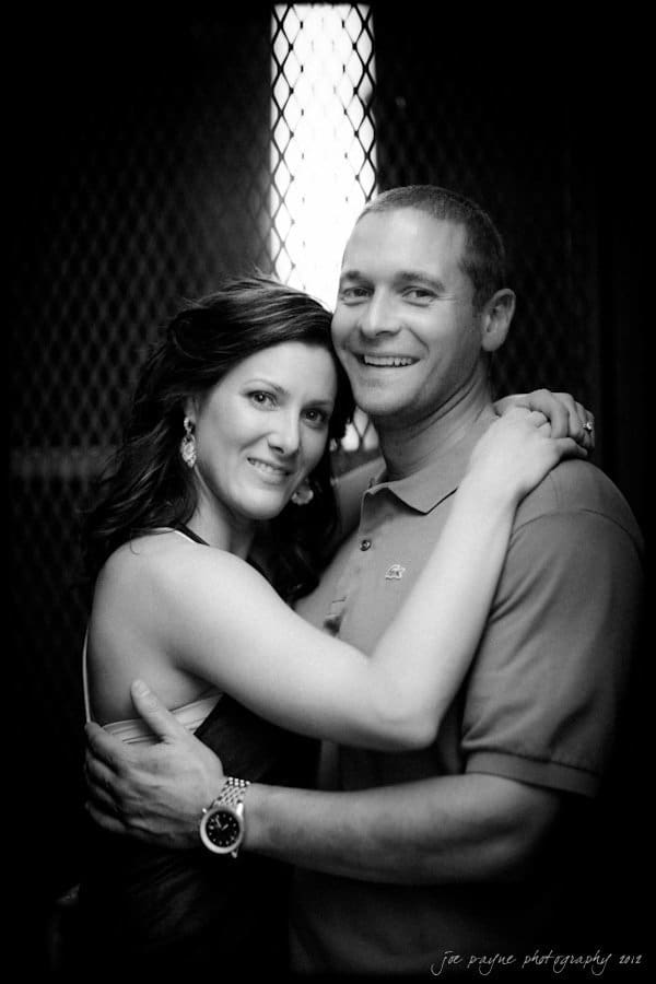 downtown raleigh engagement session ~ lisa and mark A black and white photo of a couple embracing. The woman is wearing a sleeveless dress and earrings, and the man is in a polo shirt. They are both smiling at the camera, standing in front of a dark, mesh-like background.