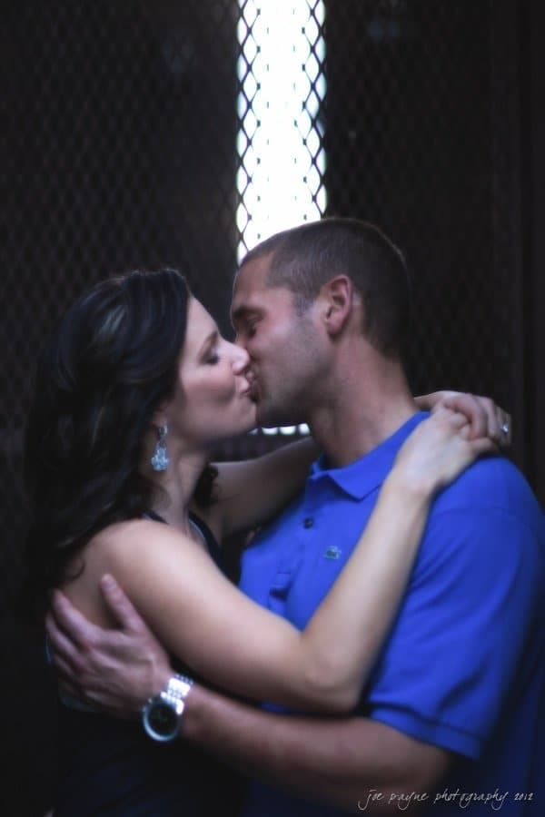 downtown raleigh engagement session ~ lisa and mark A couple embraces and kisses in a dimly lit setting. The man wears a blue shirt and the woman, with long dark hair, has her arms around his neck. They appear to be standing in front of a lattice-like background.