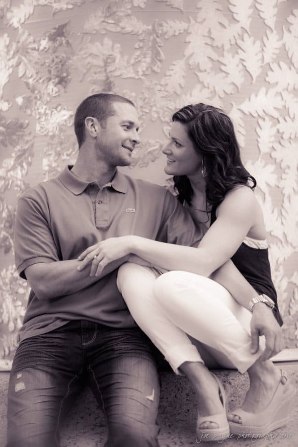 downtown raleigh engagement session ~ lisa and mark A couple sits closely together on a ledge, smiling warmly at each other. The man is in a polo shirt and jeans, and the woman is in a sleeveless top and white pants. The background features a patterned wall with leaf designs. Its a black and white image.