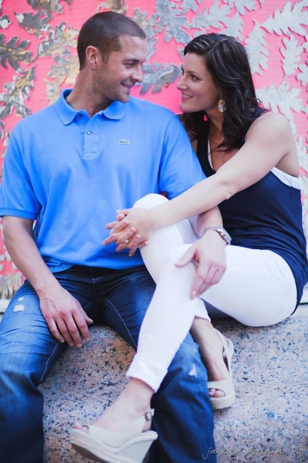 downtown raleigh engagement session ~ lisa and mark A couple sitting on a stone ledge in front of a colorful backdrop. The man wears a blue polo and jeans, while the woman is in a black top and white pants. They are looking at each other and smiling, creating a relaxed and joyful atmosphere.