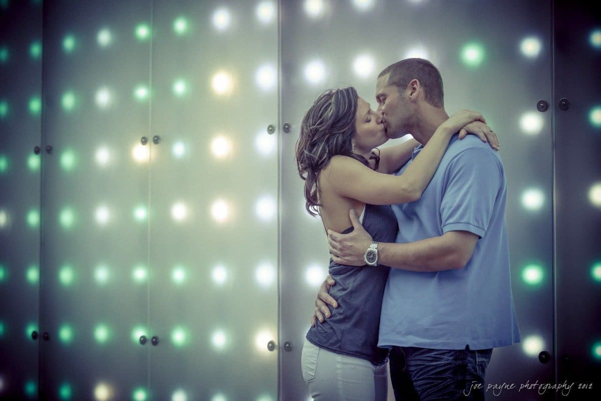downtown raleigh engagement session ~ lisa and mark A couple embraces and kisses in front of a wall illuminated with colorful lights. The background features a pattern of glowing dots in soft shades, creating a romantic and dreamy atmosphere.
