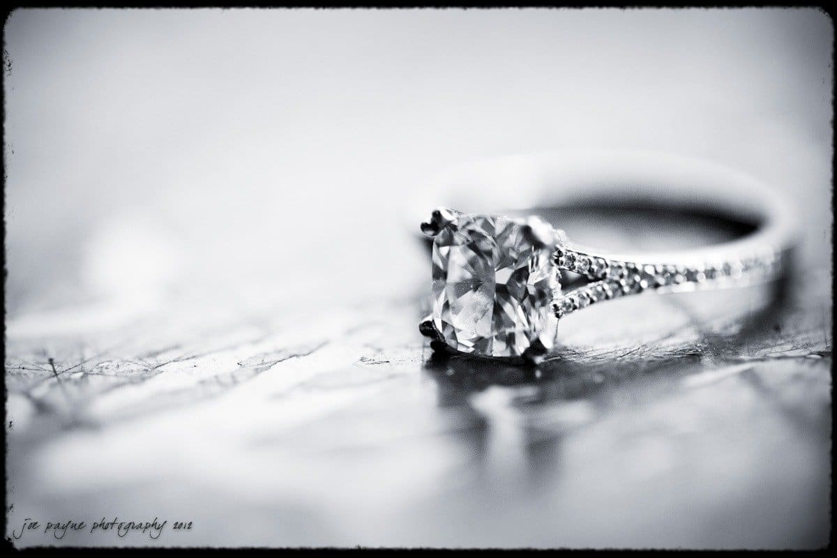 A close-up of a diamond engagement ring with a large rectangular stone and a band encrusted with smaller diamonds. The ring is placed on a textured surface, and the image has a black-and-white filter.