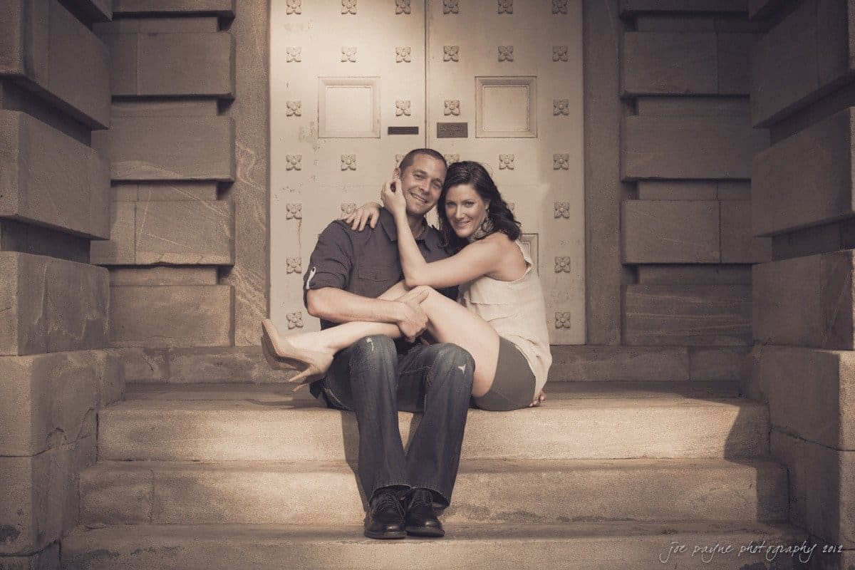 downtown raleigh engagement session ~ lisa and mark A couple sits closely together on stone steps, smiling warmly at the camera. The background features a decorative patterned door, creating an intimate and cozy atmosphere.