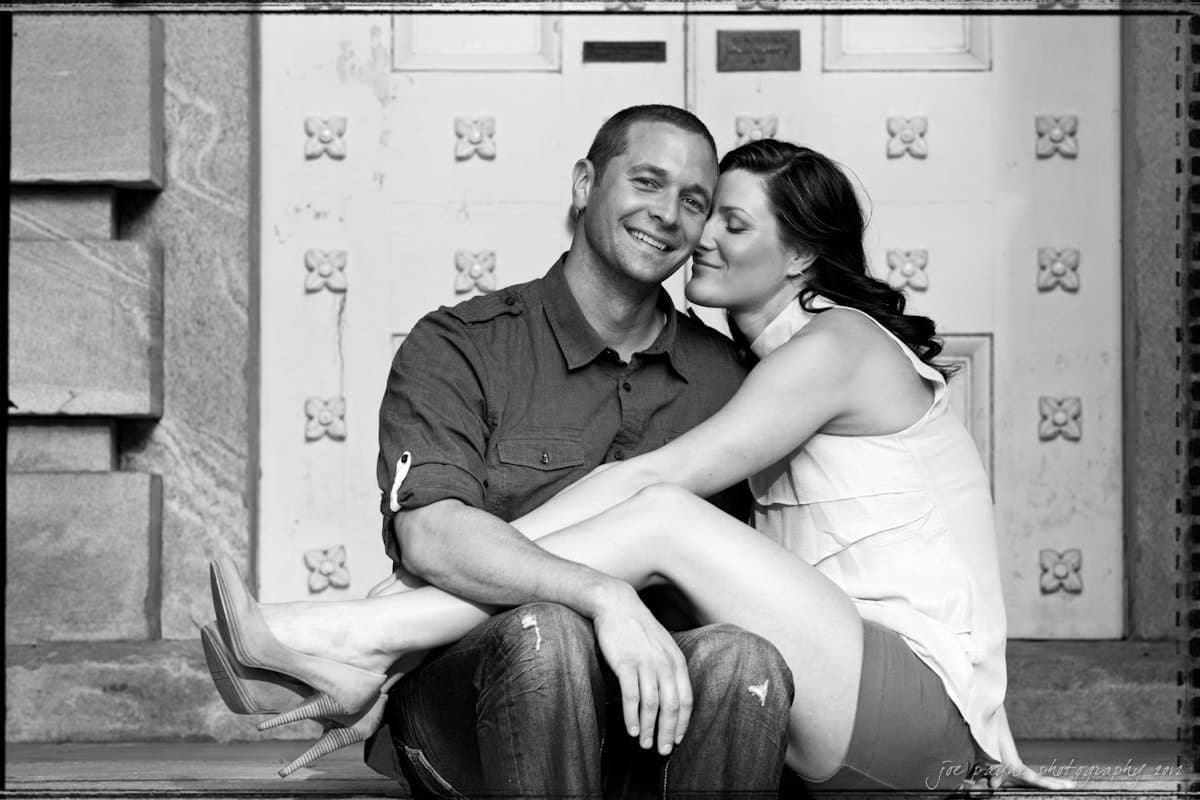 downtown raleigh engagement session ~ lisa and mark A black and white photo of a couple sitting on steps. The woman is sitting on the mans lap, leaning in with her eyes closed, smiling. The man has one arm around her waist, and they both appear content and relaxed.