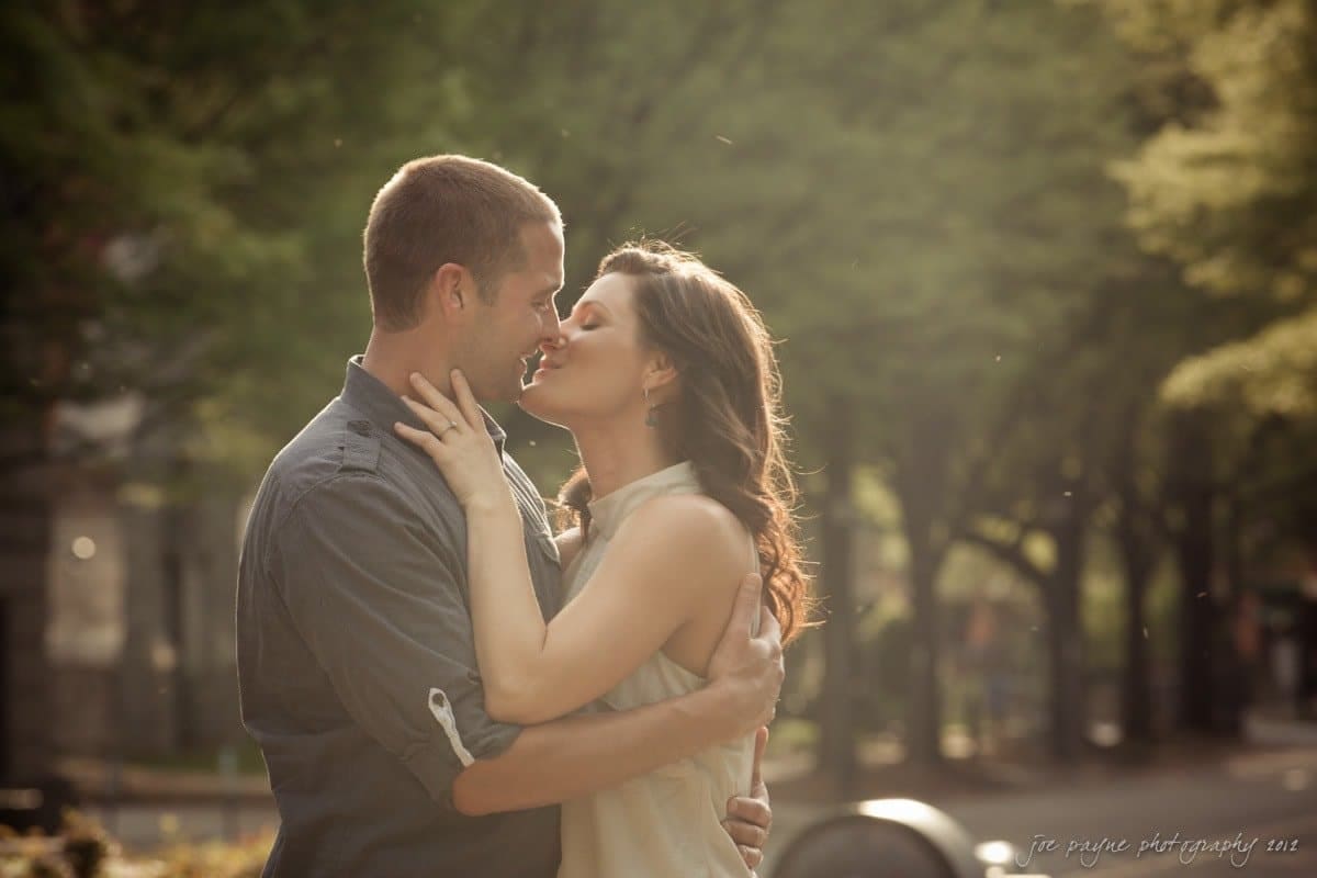 downtown raleigh engagement session ~ lisa and mark A couple embraces and shares a kiss outdoors. The background features lush green trees, and the lighting creates a soft, romantic atmosphere. The individuals appear happy and closely connected.