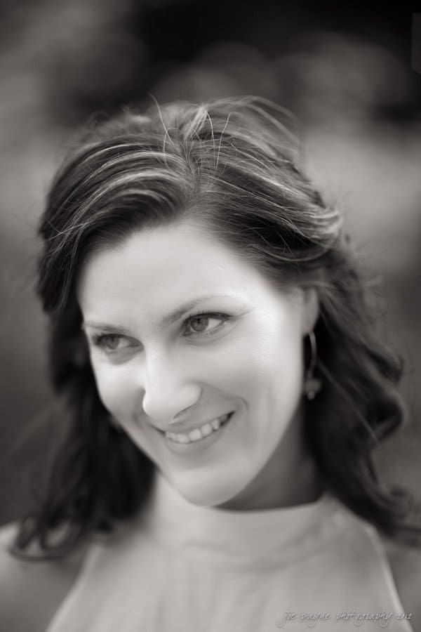 downtown raleigh engagement session ~ lisa and mark A black and white portrait of a person with long, wavy hair, smiling slightly while looking to the side. The background is blurred, highlighting the subjects face and expression.