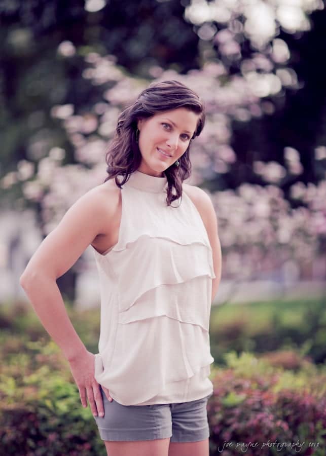 downtown raleigh engagement session ~ lisa and mark A woman with dark hair stands outdoors, wearing a sleeveless, tiered white top and gray shorts. She smiles gently, posing with hands on her hips. Trees and soft-focus flowers provide a blurred natural background.