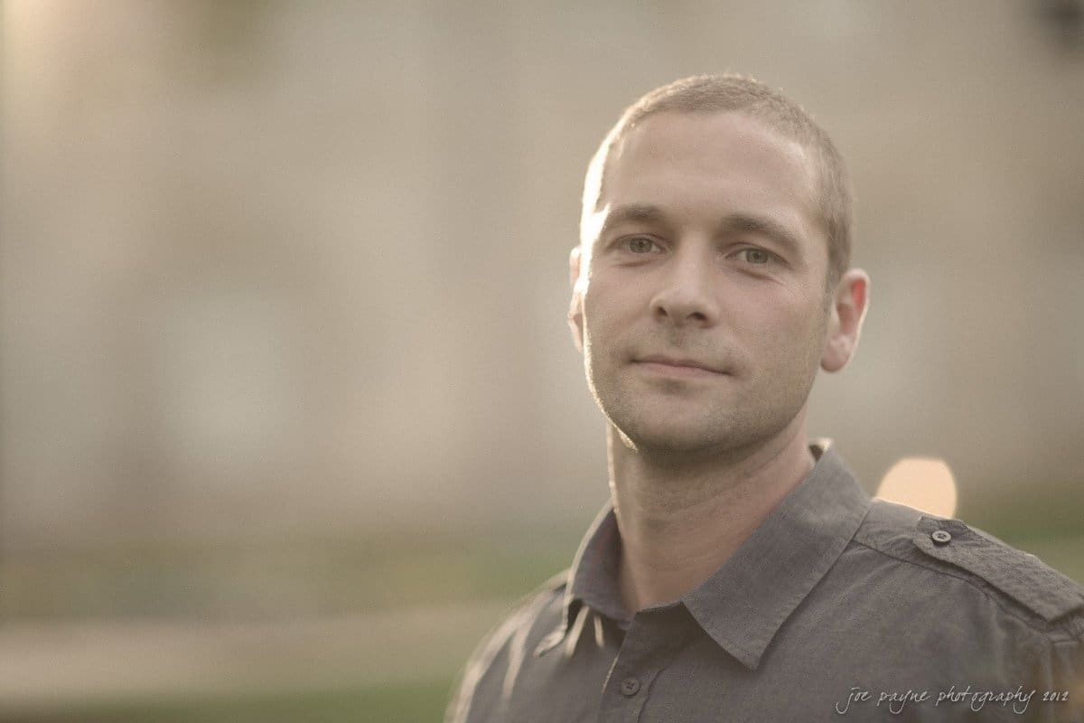 downtown raleigh engagement session ~ lisa and mark A person with short hair wearing a gray shirt smiles slightly, standing outdoors with a soft, blurred background. Sunlight gently illuminates their face, creating a warm and serene atmosphere.