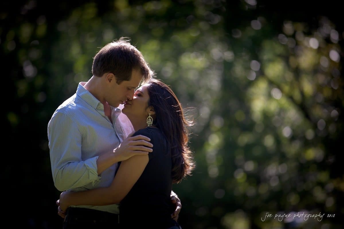 unc chapel hill engagement session ~ thelsa and phil A couple embraces affectionately in a sunlit, forested area. The man wears a light blue shirt, while the woman wears a dark top and earrings. Sunlight filters through the trees, creating a bokeh effect in the background.