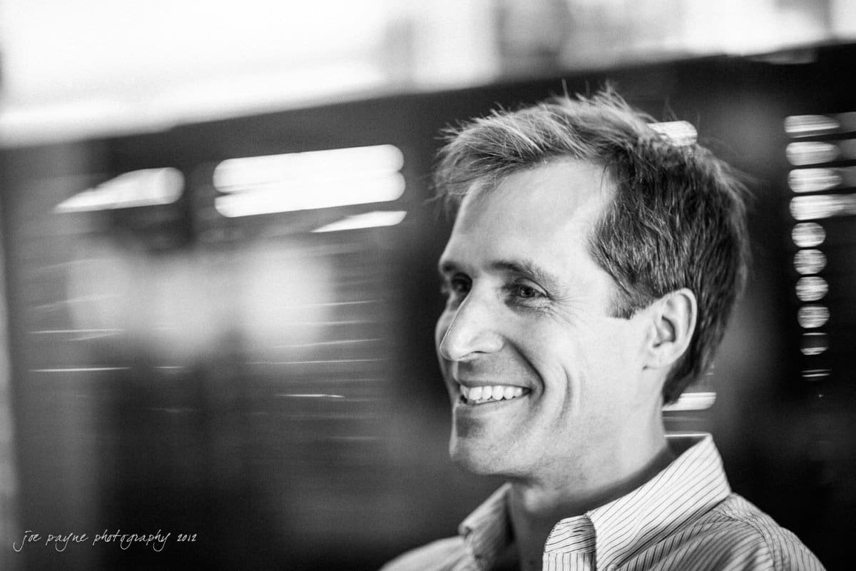 unc chapel hill engagement session ~ thelsa and phil A black-and-white photo of a smiling man wearing a striped shirt. He is looking to the right, with a blurred background. The photo has a soft focus effect.