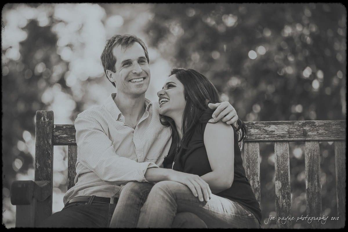 unc chapel hill engagement session ~ thelsa and phil A couple sits together on a wooden bench in a park. They are smiling and embracing, with the woman leaning towards the man. The background is a blur of trees. The image is in black and white.