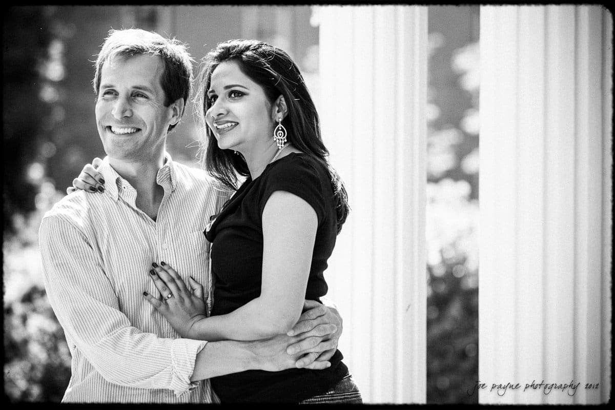 unc chapel hill engagement session ~ thelsa and phil A black and white photo of a smiling couple standing outdoors. He is wearing a striped shirt and has his arm around her. She is wearing a dark top and earrings, looking away from the camera. They stand in front of a columned structure.