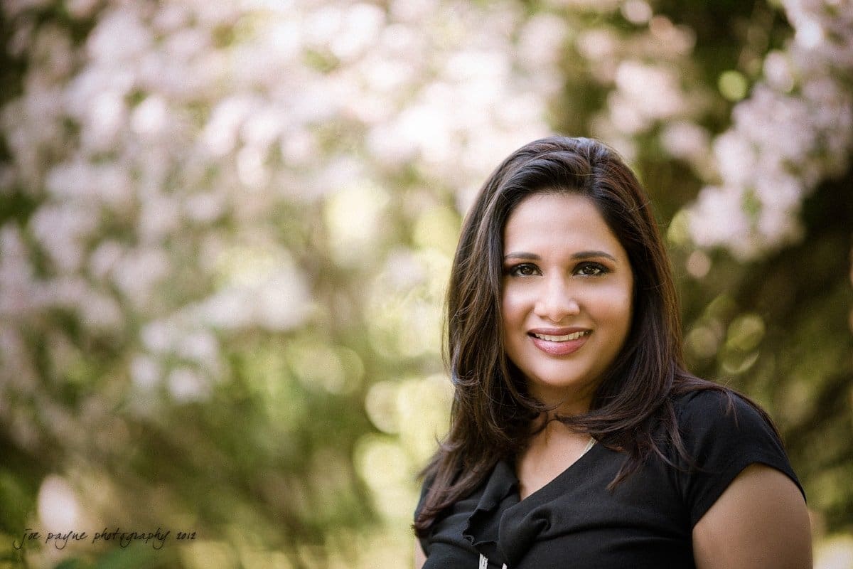 unc chapel hill engagement session ~ thelsa and phil A woman with long dark hair smiles at the camera. She is wearing a black top and standing outdoors with blurred pink flowers and greenery in the background, creating a soft, natural setting.