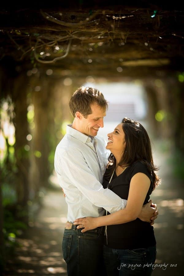 unc chapel hill engagement session ~ thelsa and phil A couple embracing and smiling at each other under a shaded, vine-covered arbor. Sunlight filters through the leaves, creating a warm and romantic atmosphere.