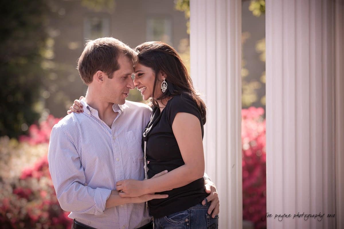 unc chapel hill engagement session ~ thelsa and phil A couple smiling and embracing near white pillars, surrounded by bright pink flowers. They appear happy and relaxed in a sunny outdoor setting.