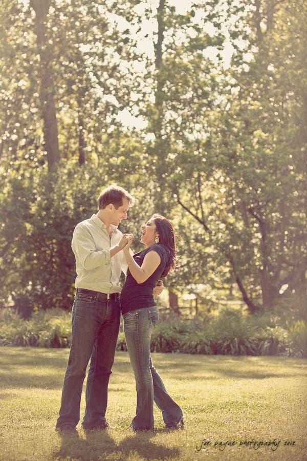 unc chapel hill engagement session ~ thelsa and phil A couple stands in a sunlit park, smiling and holding hands. Tall trees provide a green, dappled background, creating a serene and joyful atmosphere.