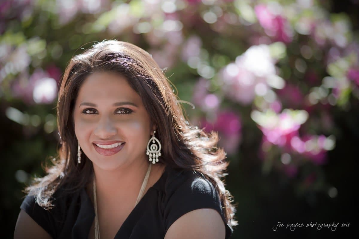 unc chapel hill engagement session ~ thelsa and phil A woman with long dark hair and dangling earrings smiles warmly while sitting outdoors. The background features vibrant pink and green blurred foliage, creating a bright and cheerful atmosphere.