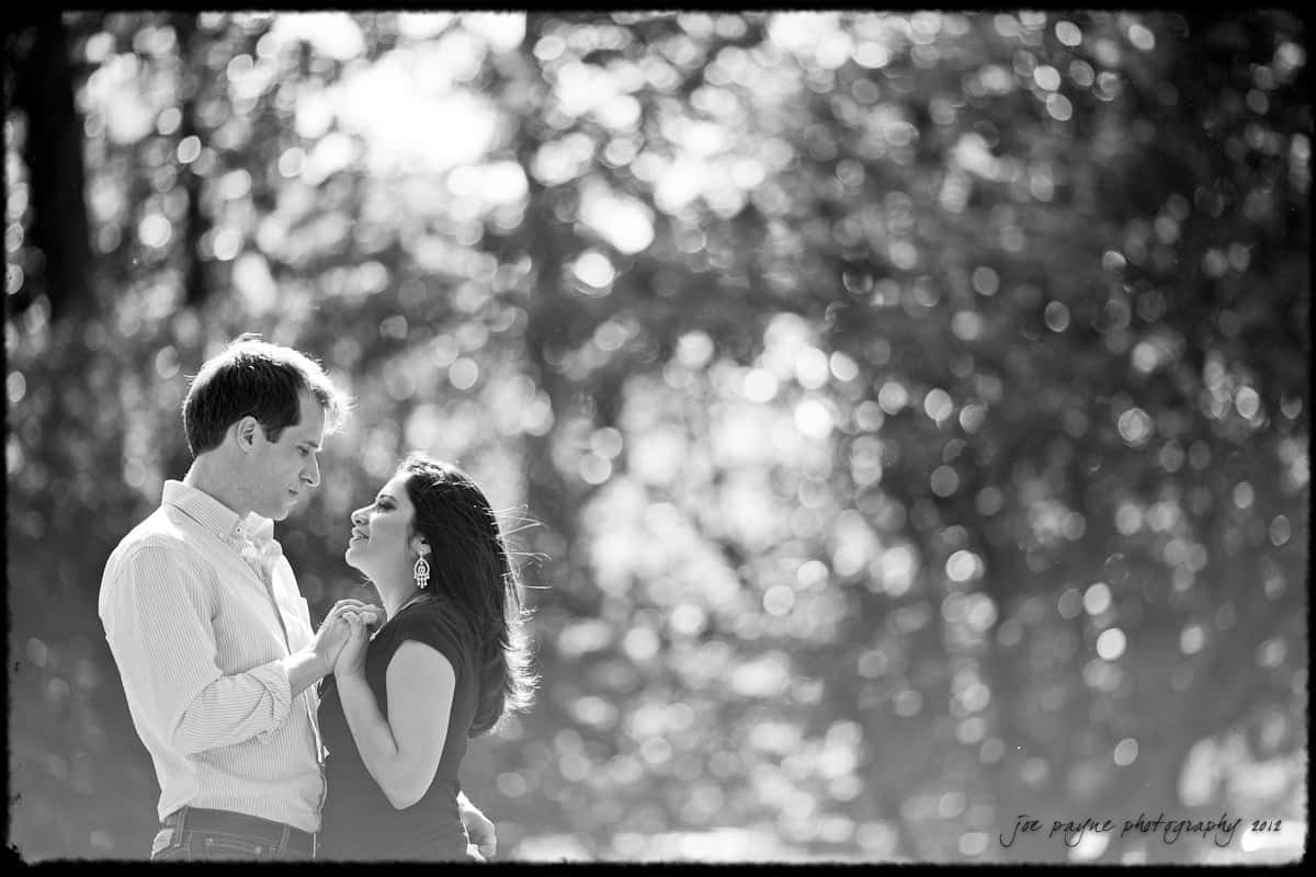 unc chapel hill engagement session ~ thelsa and phil A grayscale image of a couple gazing lovingly at each other amidst a background of blurred trees and sunlight filtering through the leaves. The woman is holding the mans hand, and both are smiling softly.