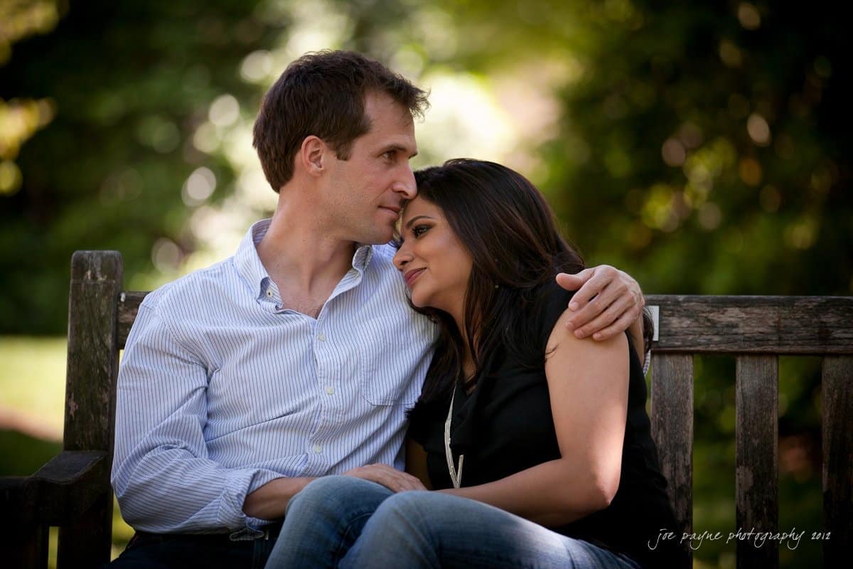 unc chapel hill engagement session ~ thelsa and phil A couple sits on a wooden bench in a park, embracing closely. The man wears a light blue shirt, and the woman wears a black top. Sunlight filters through trees in the background, creating a warm, intimate atmosphere.
