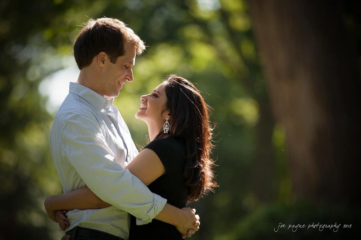 unc chapel hill engagement session ~ thelsa and phil A couple stands close in a sunlit park, smiling at each other. The man in a striped shirt embraces the woman in a black dress, who looks back at him warmly. Lush greenery is softly blurred in the background.