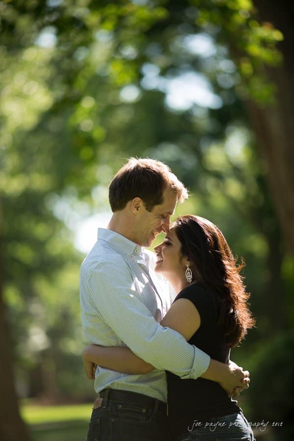 unc chapel hill engagement session ~ thelsa and phil A couple embraces and smiles at each other in a sunlit, green park. They are surrounded by blurred trees and sunlight filtering through the leaves, creating a romantic atmosphere.