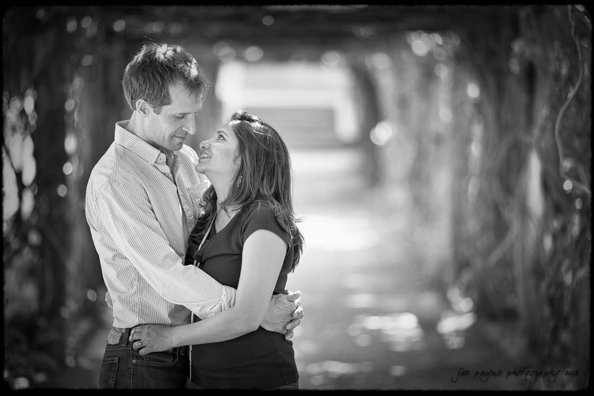unc chapel hill engagement session ~ thelsa and phil A black and white photo of a couple embracing and smiling at each other in a sunlit, leafy walkway. The background is softly blurred, highlighting the couples affectionate gaze.