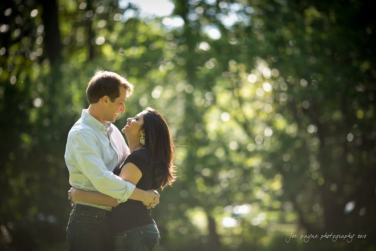 unc chapel hill engagement session ~ thelsa and phil A couple embraces and smiles at each other in a sun-dappled forest setting. The background is lush and green, with light filtering through the leaves, creating a soft, romantic atmosphere.