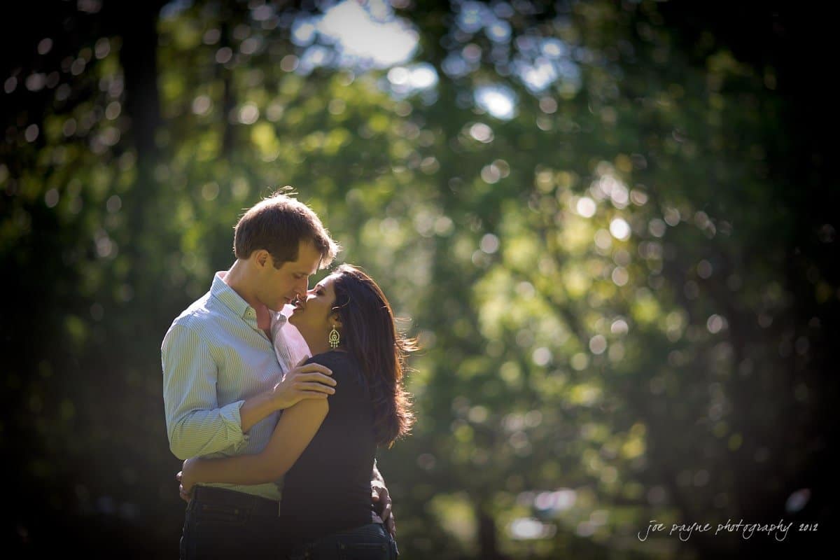 unc chapel hill engagement session ~ thelsa and phil A couple embraces lovingly in a sun-dappled, leafy park. The man wears a light blue shirt, while the woman sports a dark top and hoop earrings. Sunlight filters through the trees, creating a serene and intimate atmosphere.
